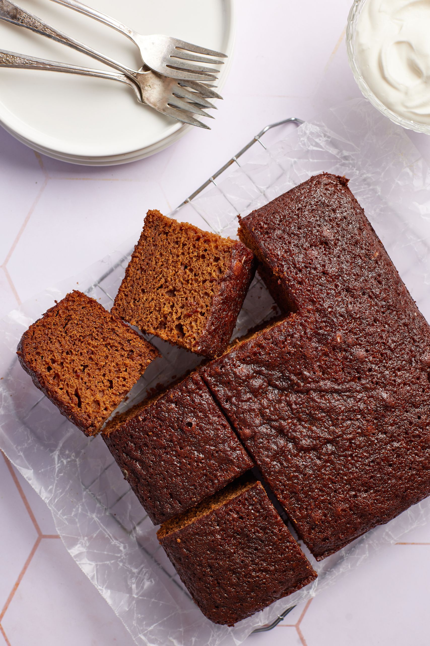 A molasses cake has been cut into 9 big squares on the cooling rack. Two are having the sides facing up, showing the moist and airy crumb. Whipped cream is served on the side. 