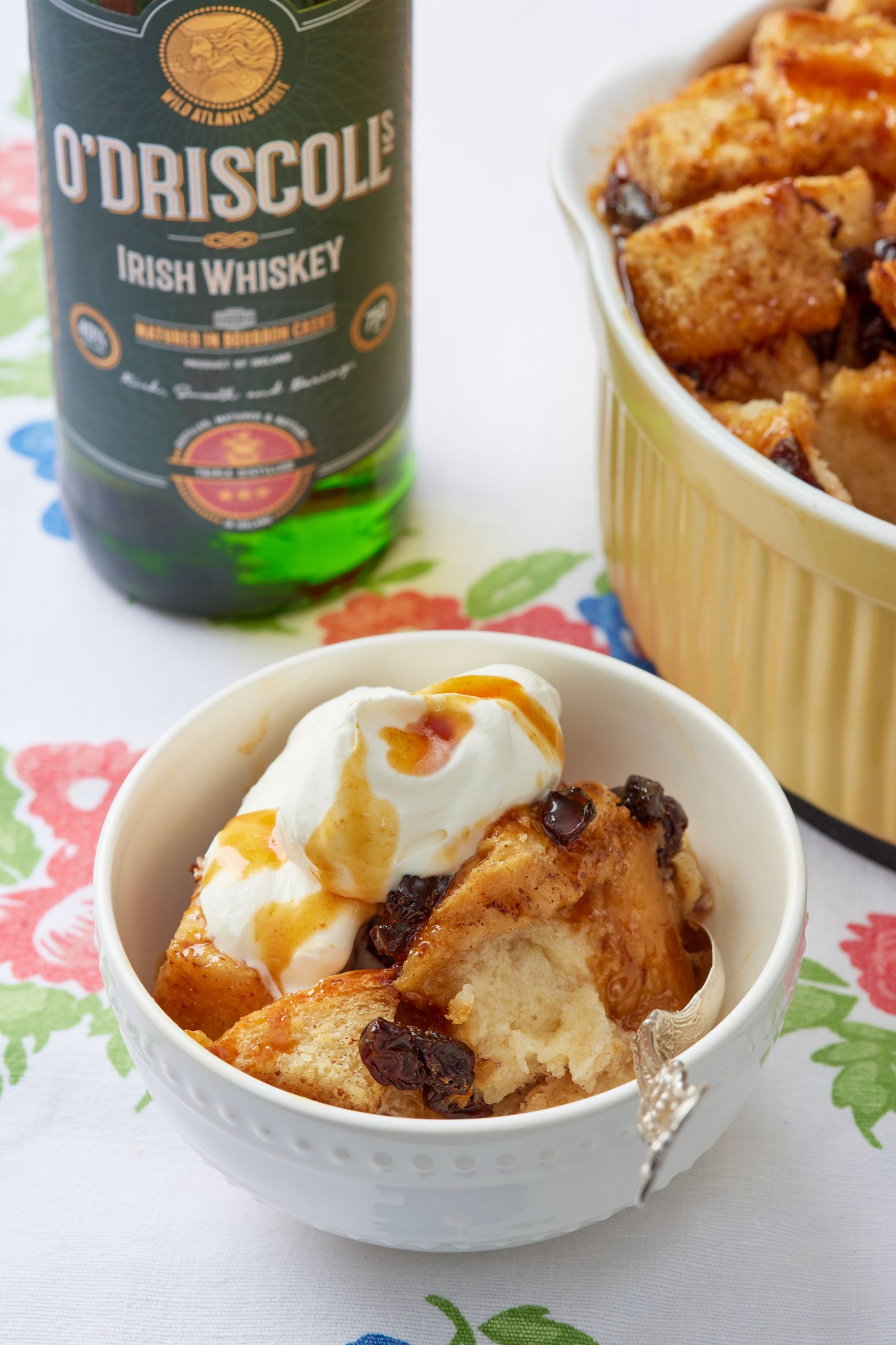 A close-up shot at a dessert bowl of bread pudding served with softly whipped cream and whiskey sauce. A bottle of Irish Whiskey and a bowl of the remaining pudding are in the background. 