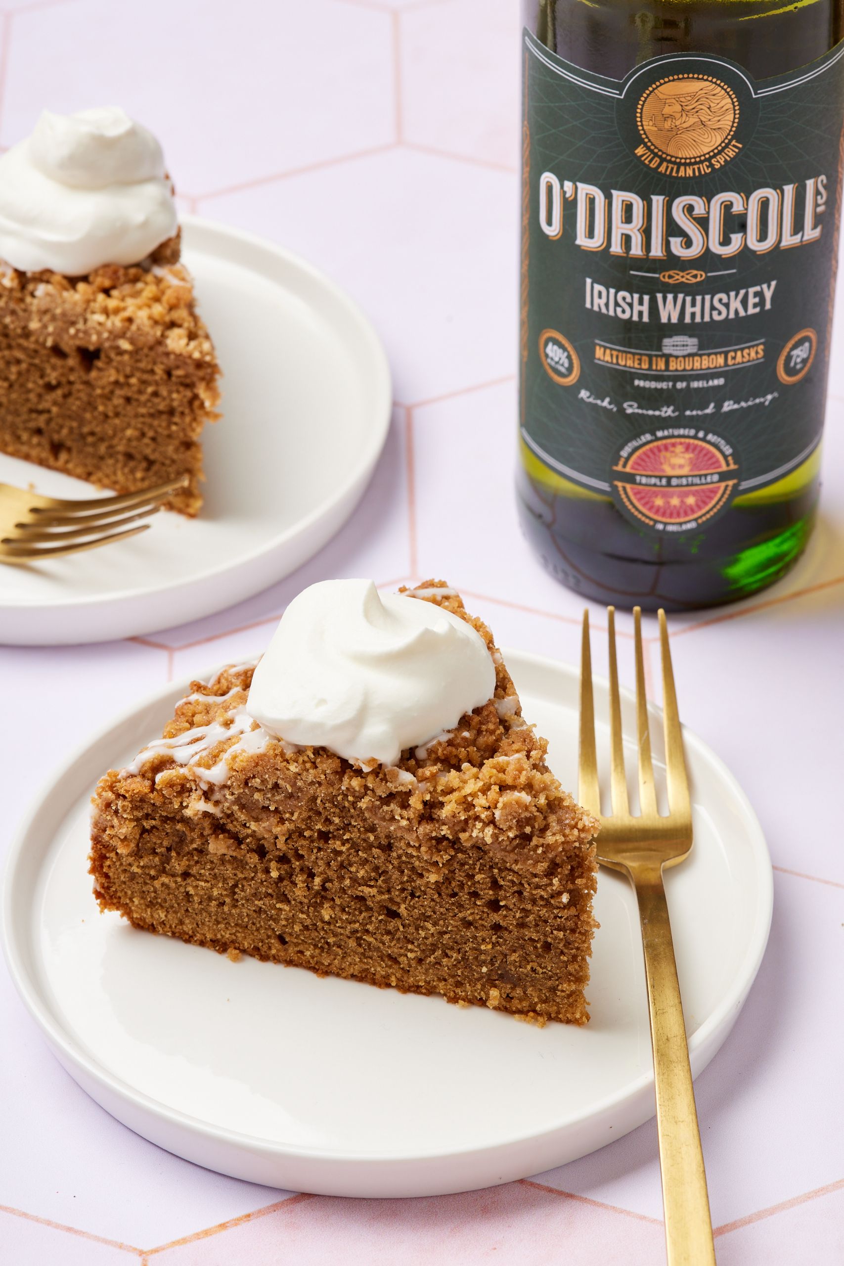 A close-up shot at two slices of Irish Coffee Cake topped with softly whipped cream, with a bottle of Irish whiskey on the side. 
