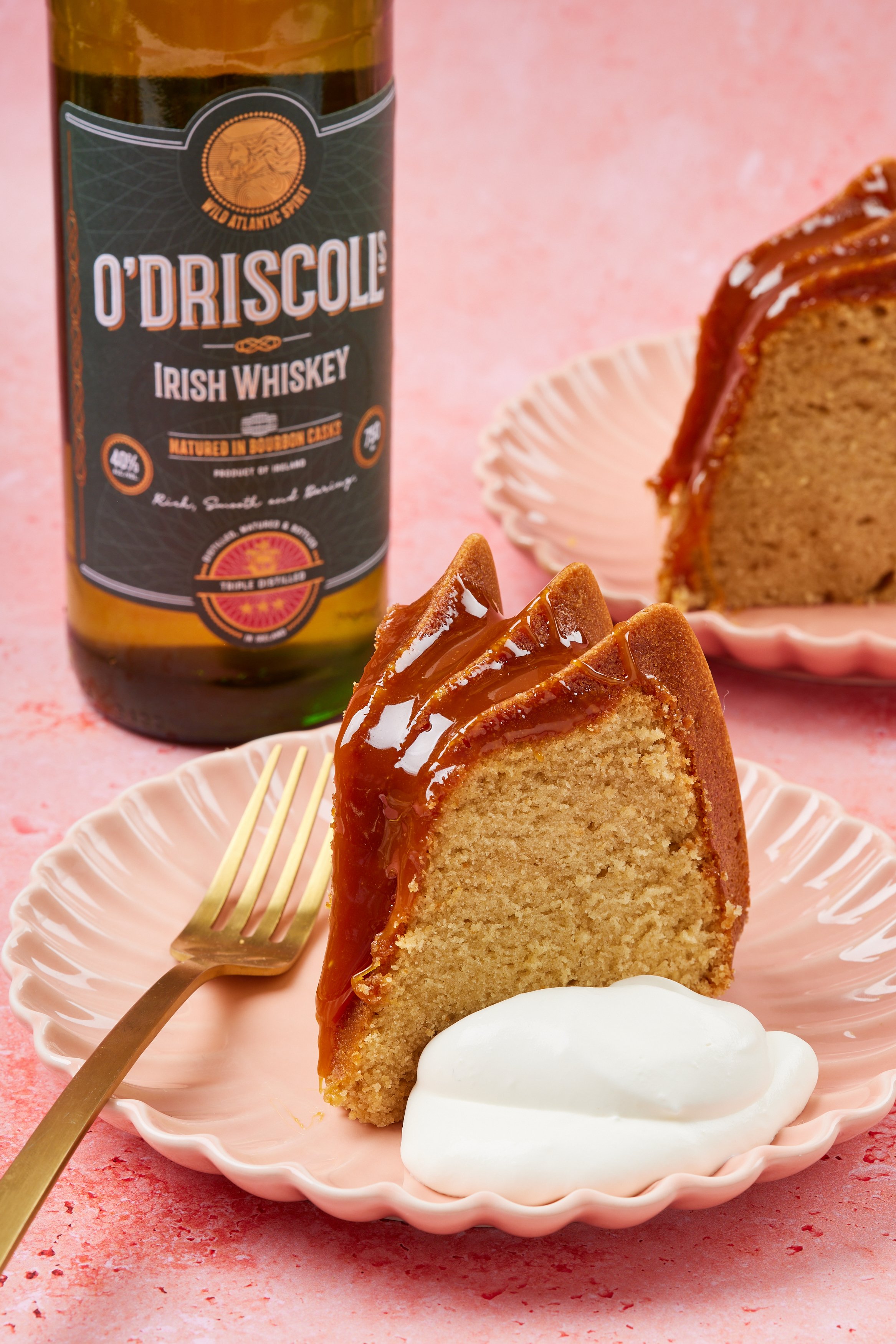 A close-up shot at a slice of Whiskey Cake drizzled with whiskey caramel sauce served with softly whipped cream. A bottle of Irish Whiskey and a second slice of cake are in the back. 
