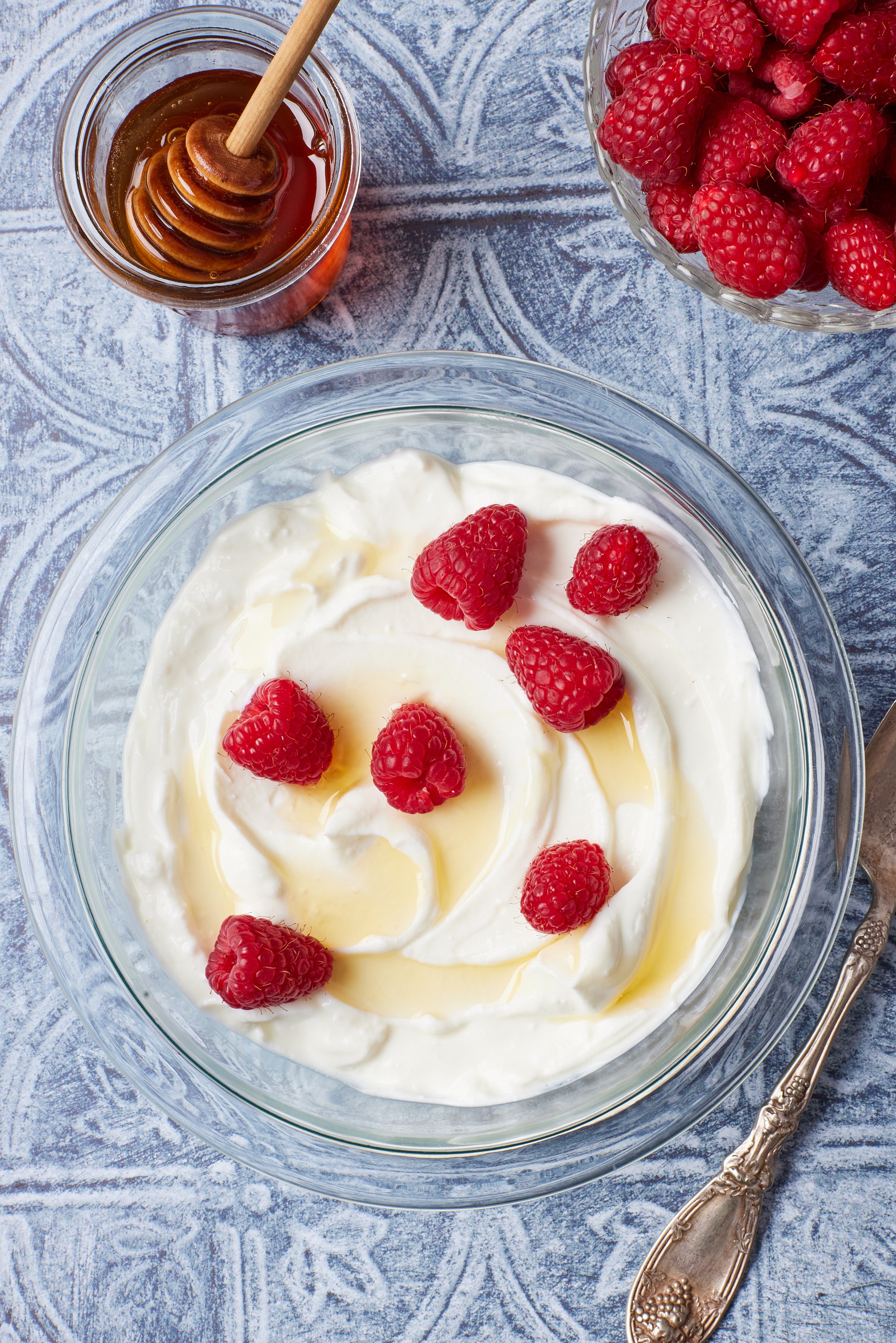 Greek Yogurt12 Top down view of a bowl of homemade yogurt topped with fresh raspberries, with more raspberries and honey on the side .