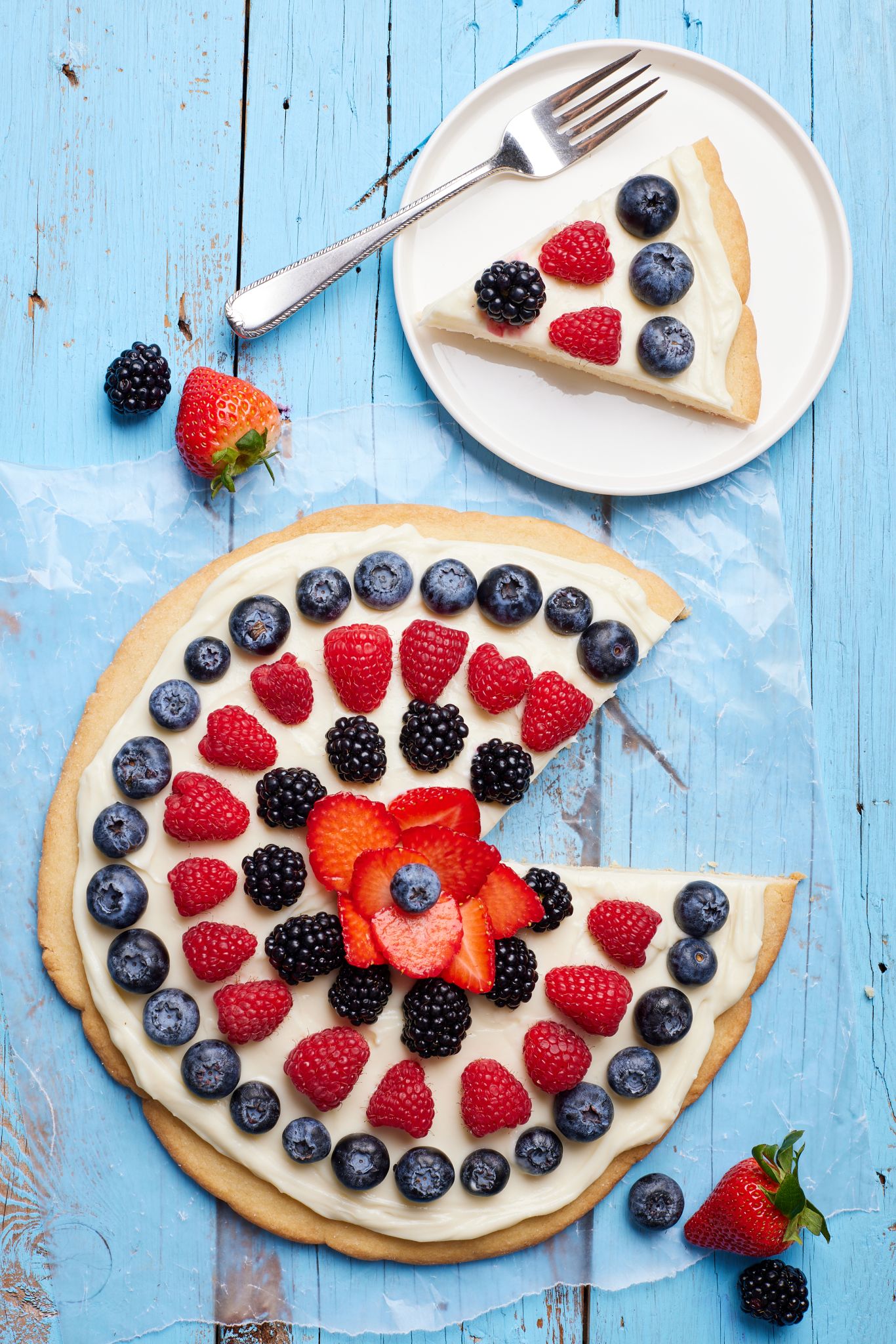 A top-down view of the July 4th Fruit Pizza, with one slice cut and served on a dessert plate. 