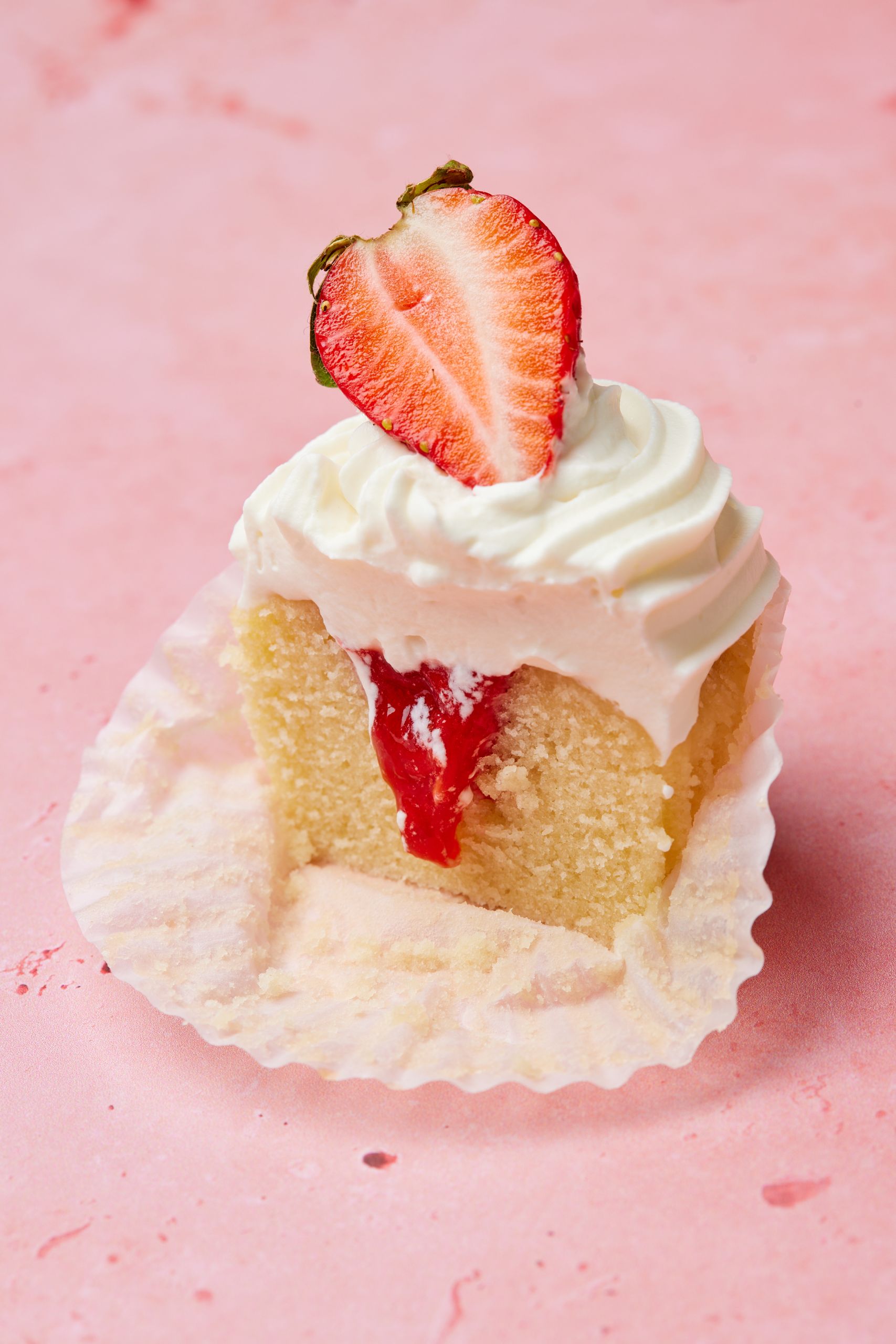 A close-up shot at a sliced cupcake shows the moist cake with compote filling, topped with silky frosting and fresh strawberry slice. 