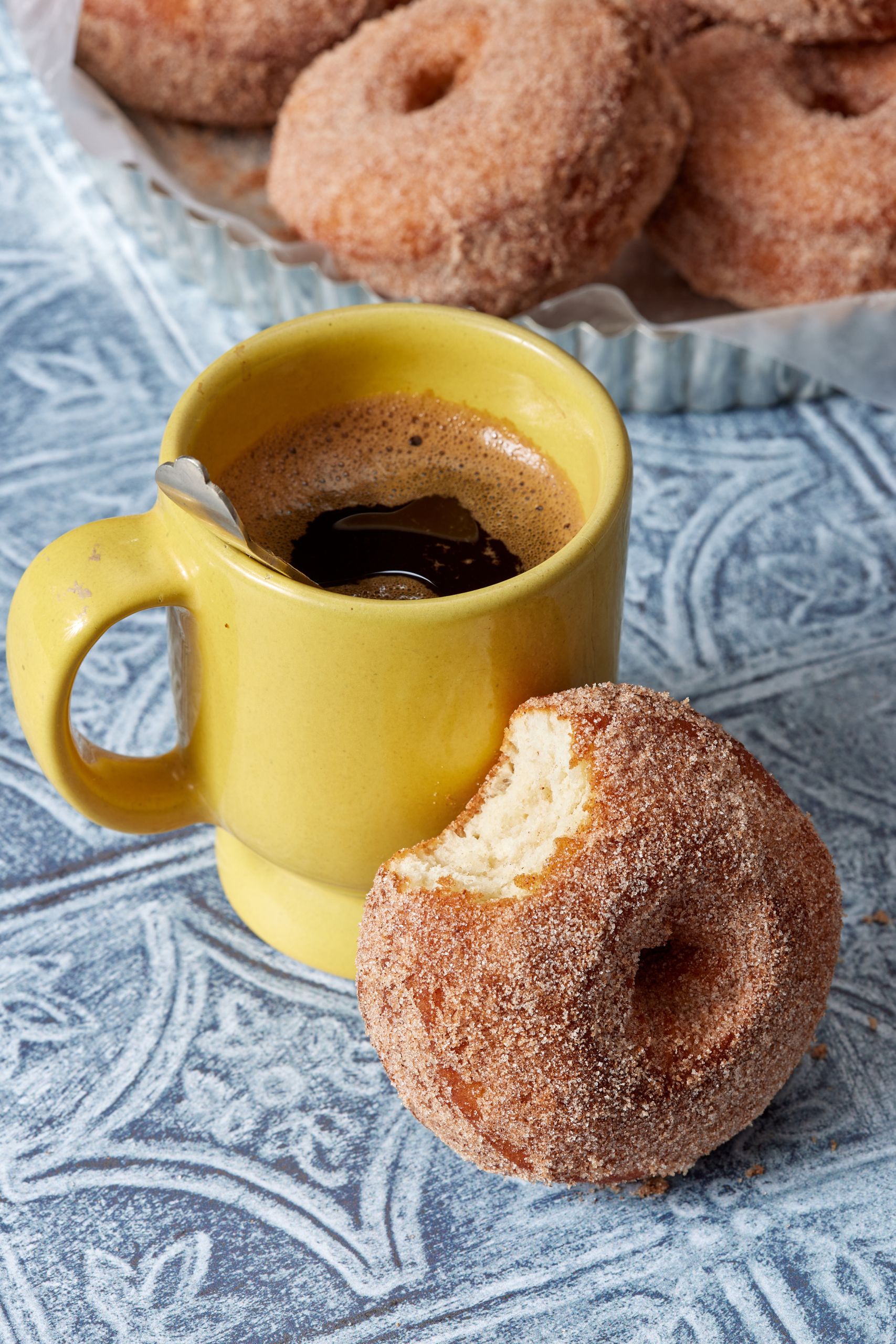A potato donut with one bite taken with a cup of coffee.