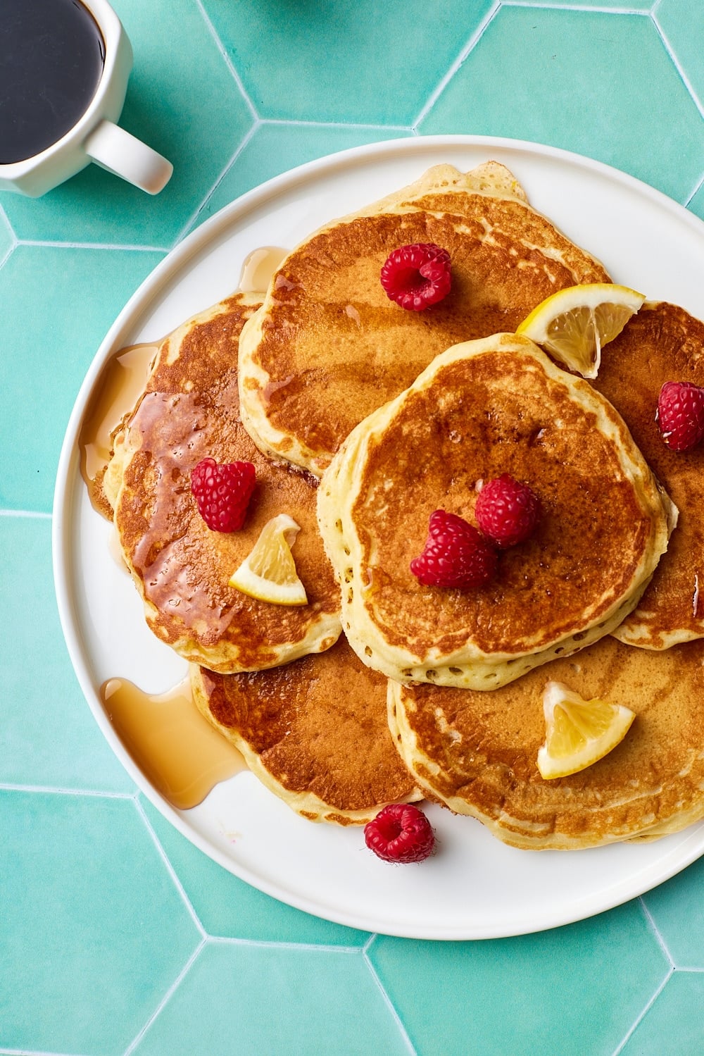 Top down view of a plate of golden fluffy lemon pancakes served with fresh raspberries and syrup, with coffee on the side. 