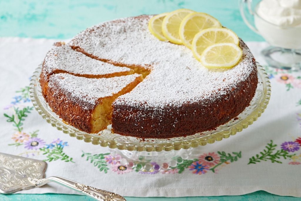 A Lemon Torta Caprese cake on the stand is dusted with powder sugar and decorated with lemon slices on top.