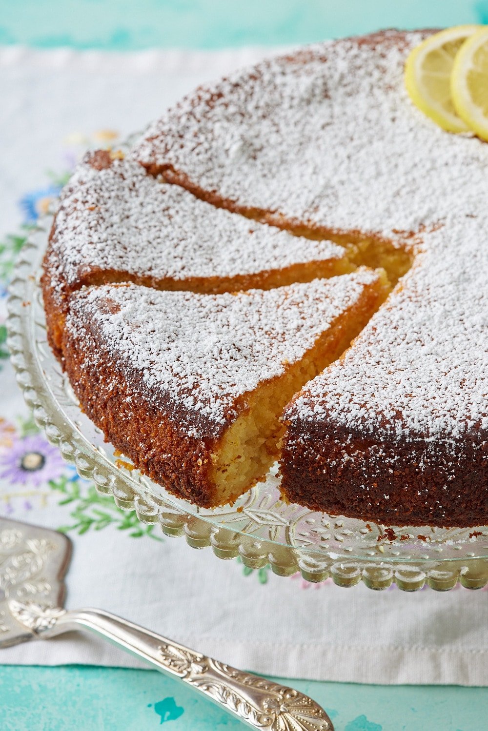 Torta Caprese Bianca9 A Lemon Torta Caprese cake on the stand is dusted with powder sugar and decorated with lemon slices on top. Two slices are cut.