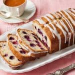 A glazed loaf of Lemon Blueberry Bread. Part of it is sliced and shows the moist crumb. Served with tea on the side.