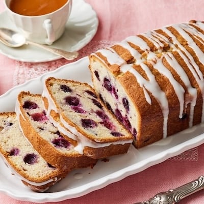 A glazed loaf of Lemon Blueberry Bread. Part of it is sliced and shows the moist crumb. Served with tea on the side.