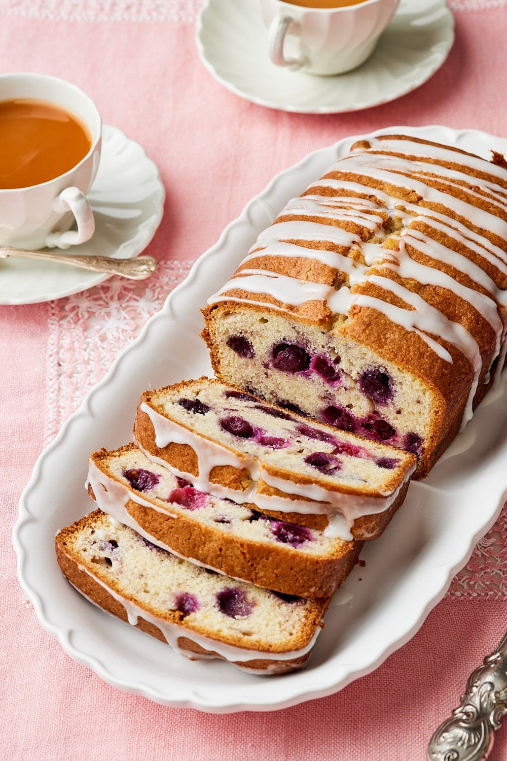 Lemon blueberry bread7 A glazed loaf of Lemon Blueberry Bread. Part of it is sliced and shows the moist crumb. Served with tea on the side.