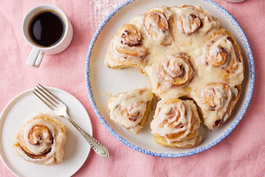 Top-dwon view of Sourdough Cinnamon rolls with cream cheese glaze. One roll is served separately on a plate and served with coffee.