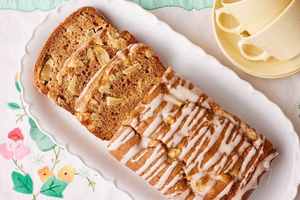 Whole Apple Loaf Cake drizzled with apple glaze and sliced to reveal a moist cinnamon-spiced crumb filled with diced apples on a white serving platter.