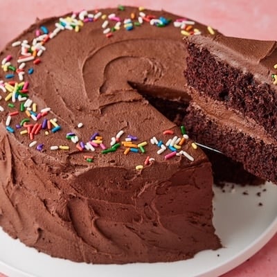 Chocolate Mayonnaise Cake decorated with sprinkles on top. Oe slice being cut, showing the moist crumb and layers of rich buttercream frosting.