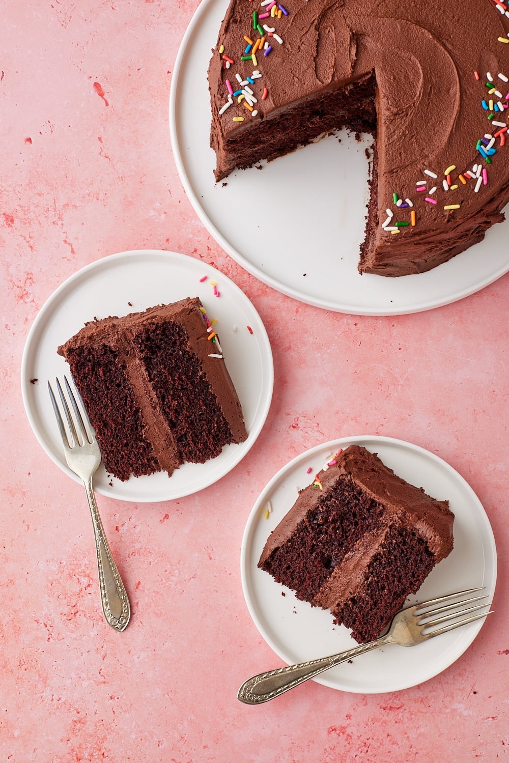 Chocolate Mayo Cake15.1 Overhead view of a Chocolate Mayonnaise Cake frosted with chocolate buttercream and decorated with rainbow sprinkles, with two slices served on white dessert plates alongside forks.