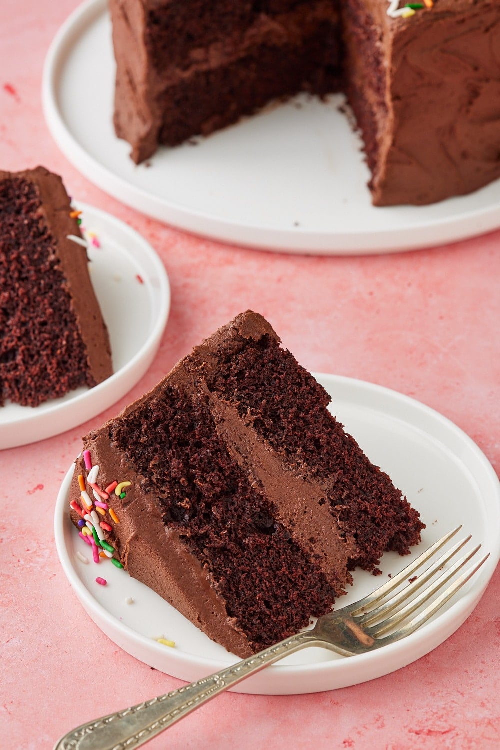 Chocolate Mayo Cake15 Slice of Chocolate Mayonnaise Cake on a white dessert plate with a fork, showing moist chocolate layers filled and frosted with creamy chocolate buttercream, with the remaining cake in the background.