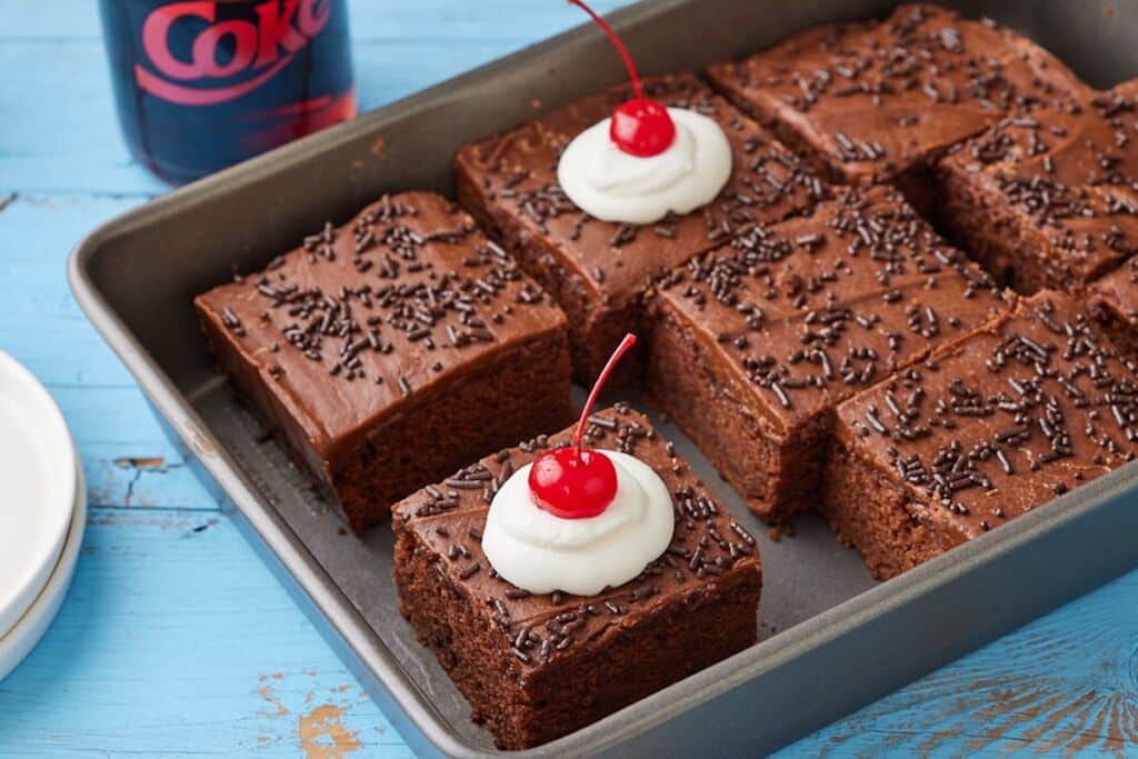 Coca Cola Cake squares in a baking tray, with moist crumb, rich frosting, whipped cream and cherries.