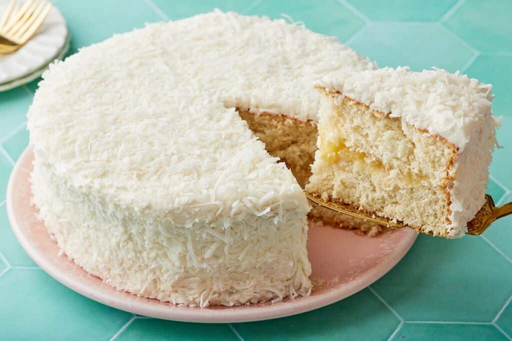 Pineapple Coconut Cake coated with shredded coconut, with one slice cut, showing the moist crumbs.