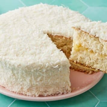 Pineapple Coconut Cake coated with shredded coconut, with one slice cut, showing the moist crumbs.