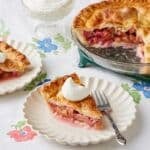 Two slices of rhubarb pie with golden crust and bubbly filling are cut and served next to the remaining pie, with whipped cream on top.