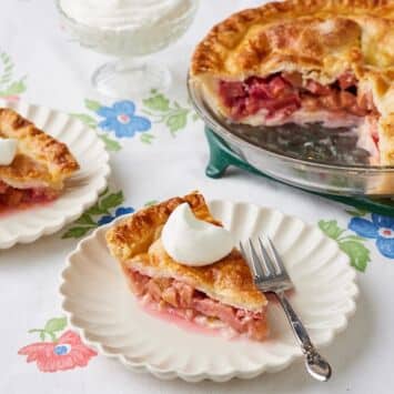 Two slices of rhubarb pie with golden crust and bubbly filling are cut and served next to the remaining pie, with whipped cream on top.