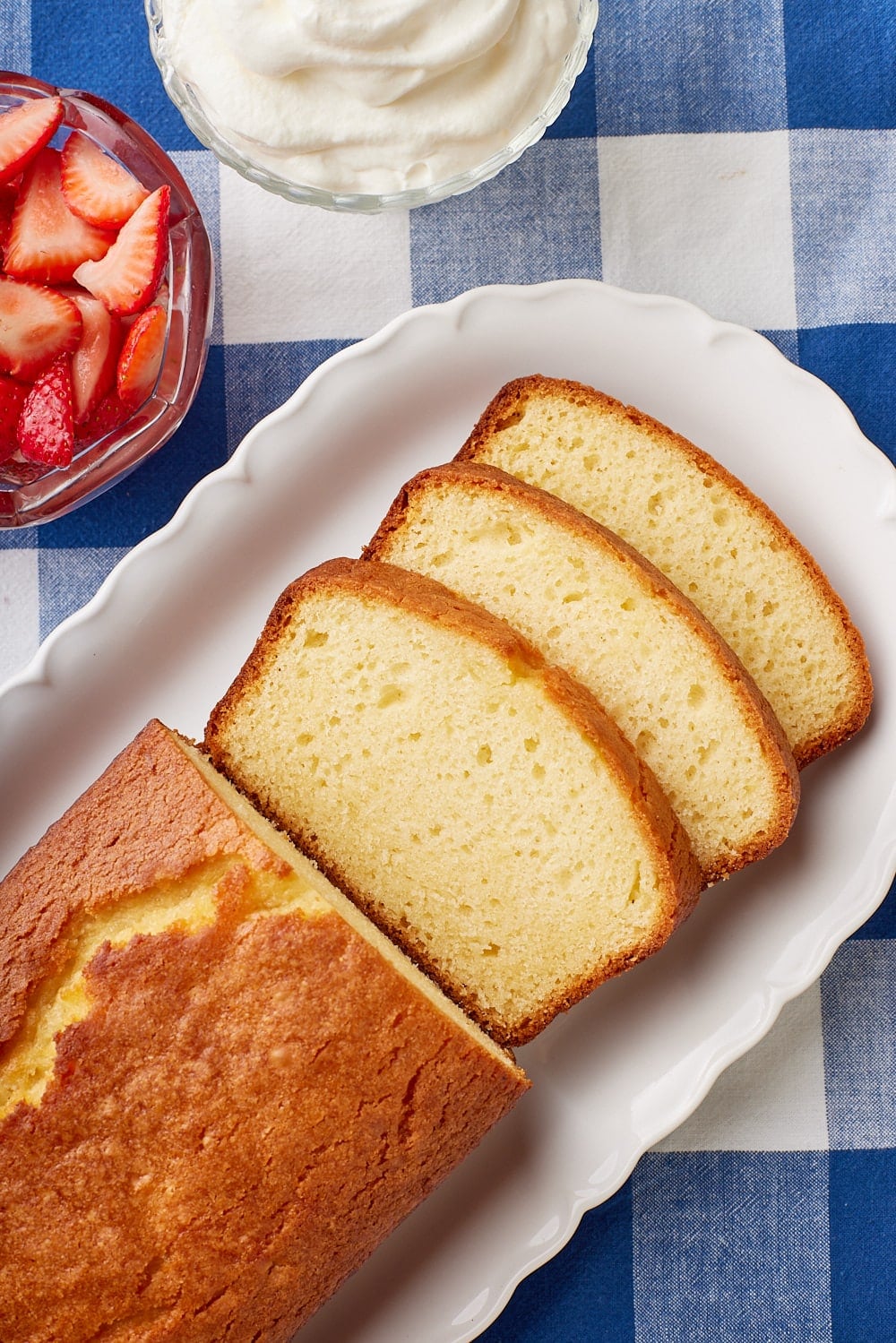 Top-down view of French Yogurt Cake on a platter, with half of it sliced. Fresh strawberry slices and whipped cream are served on the side. 