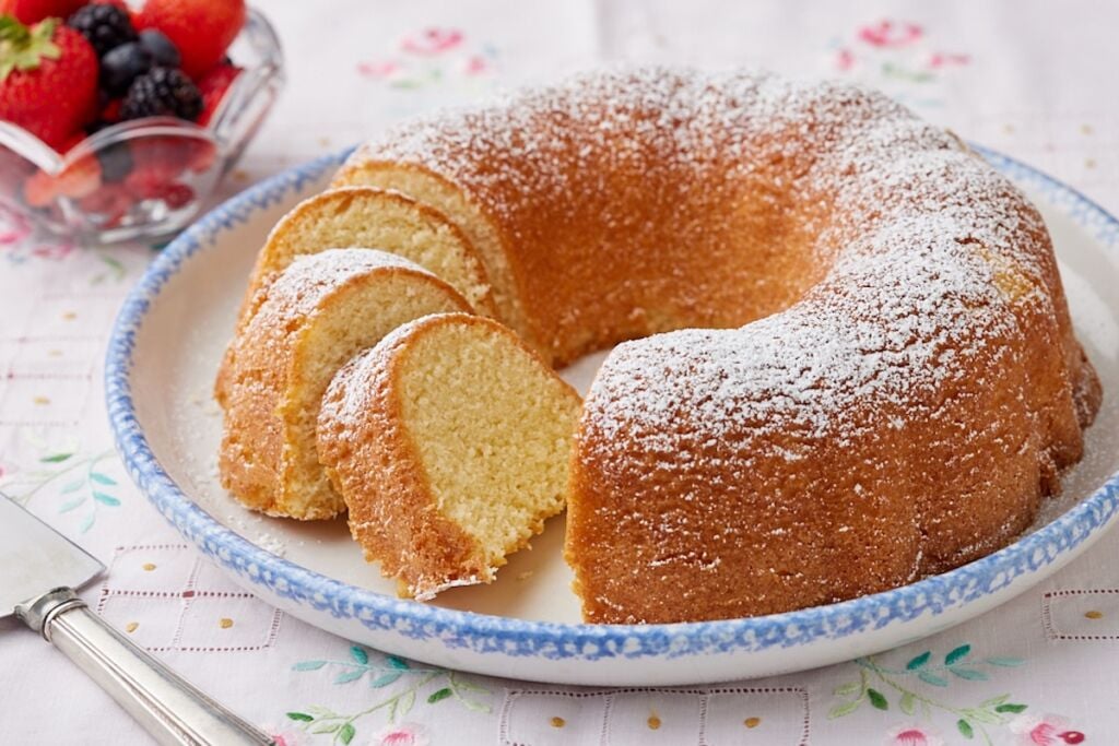 A Hot Milk Cake is dusted with powdered sugar and partially sliced, served with fresh berries on the side.