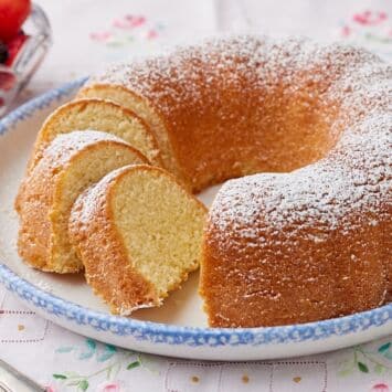 A Hot Milk Cake is dusted with powdered sugar and partially sliced, served with fresh berries on the side.