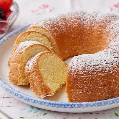 A Hot Milk Cake is dusted with powdered sugar and partially sliced, served with fresh berries on the side.