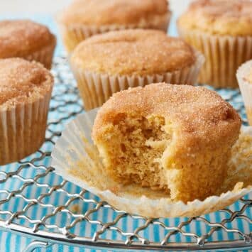 Apple cider muffins with cinnamon-sugar tops, one muffin split open to show its soft, tender crumb, arranged on a wire cooling rack.