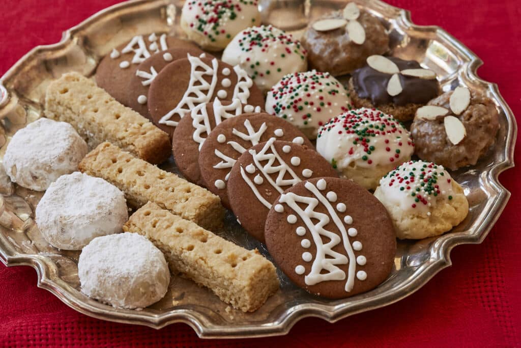 A festive European Christmas cookie platter featuring an assortment of gingerbread cookies, shortbread, powdered sugar cookies, iced cookies with red and green sprinkles, and almond-topped cookies arranged on a silver serving tray over a red tablecloth.
