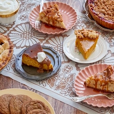 A variety of homemade holiday pies and desserts arranged on a lace-covered table, including apple pie, pumpkin pie, cherry crumble pie, cheesecake, pumpkin cake, and cookies.