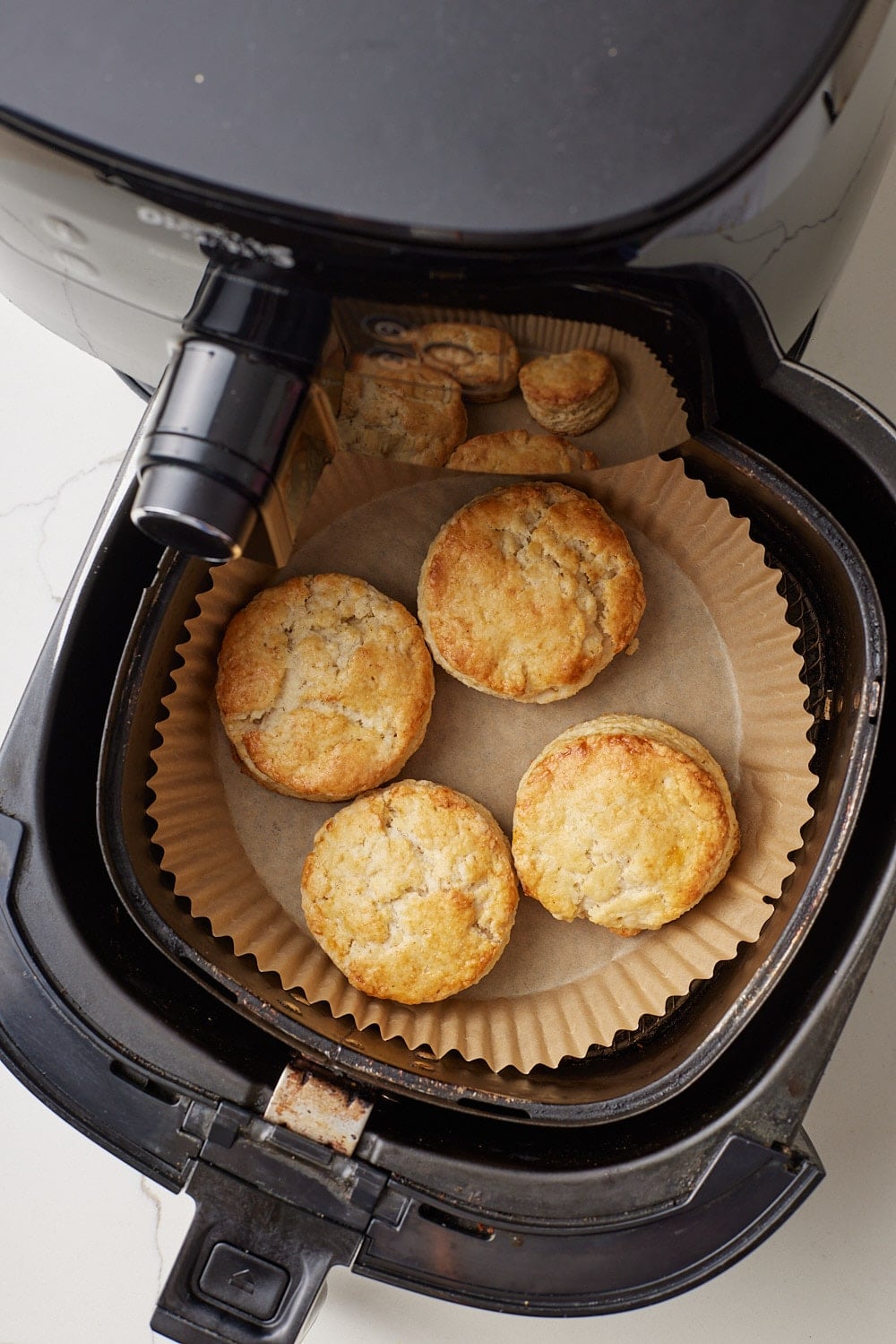 Air Fryer Scones7 Four golden Air Fryer Scones cooking in a parchment-lined air fryer basket, with another batch visible inside the appliance.