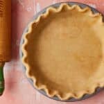 Overhead view of homemade brown butter pie crust pressed into a round pie pan with crimped edges, alongside a wooden rolling pin on a pink surface.