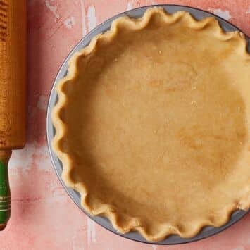 Overhead view of homemade brown butter pie crust pressed into a round pie pan with crimped edges, alongside a wooden rolling pin on a pink surface.