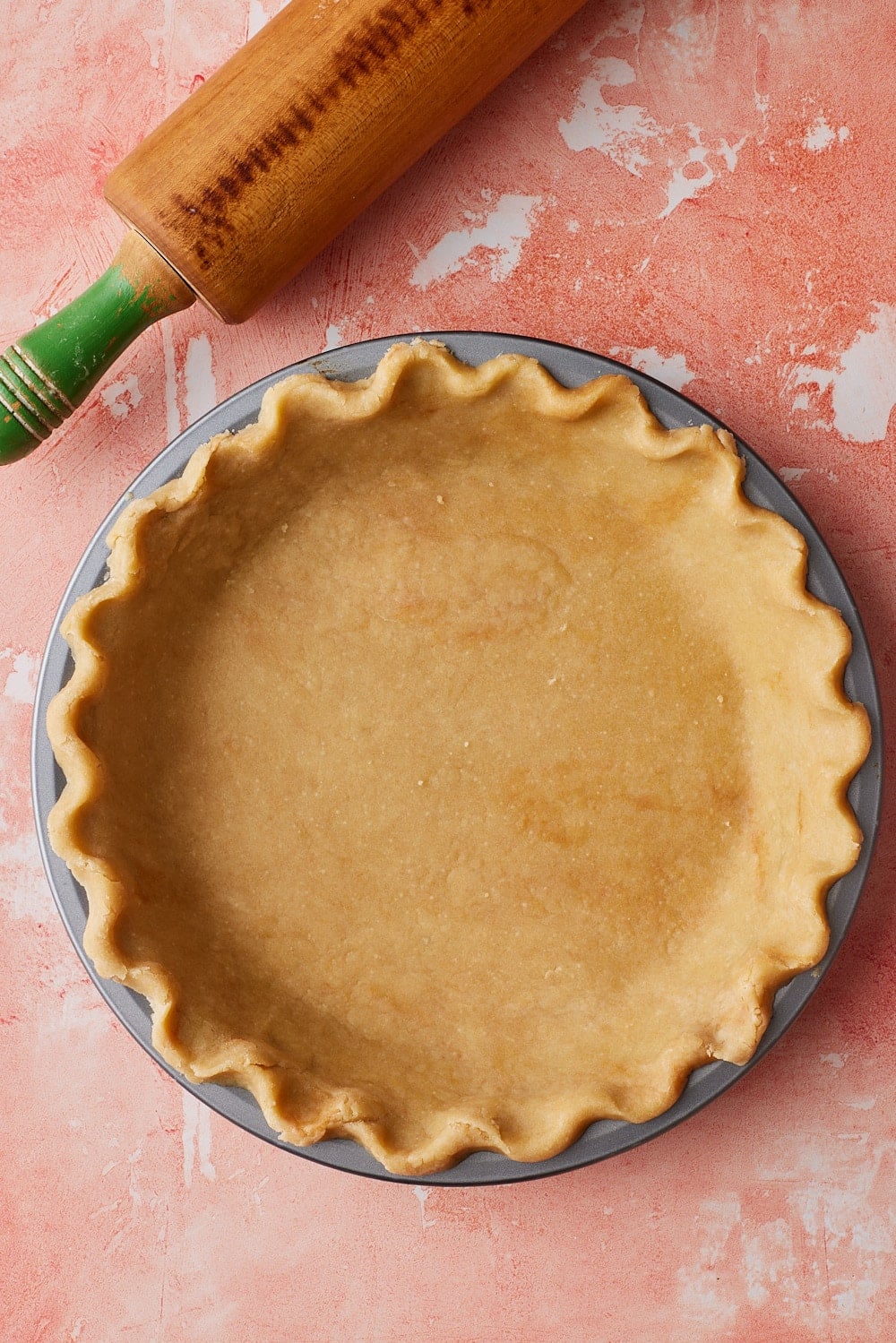 Overhead view of homemade brown butter pie crust fitted into a round pie pan with decorative crimped edges, alongside a wooden rolling pin on a pink countertop.