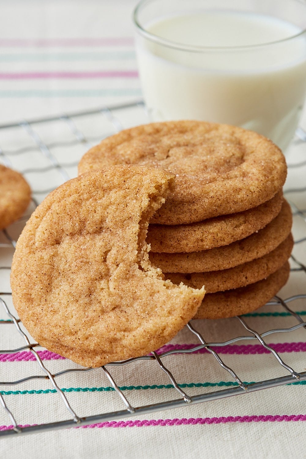 Brown Butter Snickerdoodles11 Stacked brown butter snickerdoodle cookies coated in cinnamon sugar, with one cookie broken open to show its soft, chewy center, arranged on a wire cooling rack with a glass of milk in the background.
