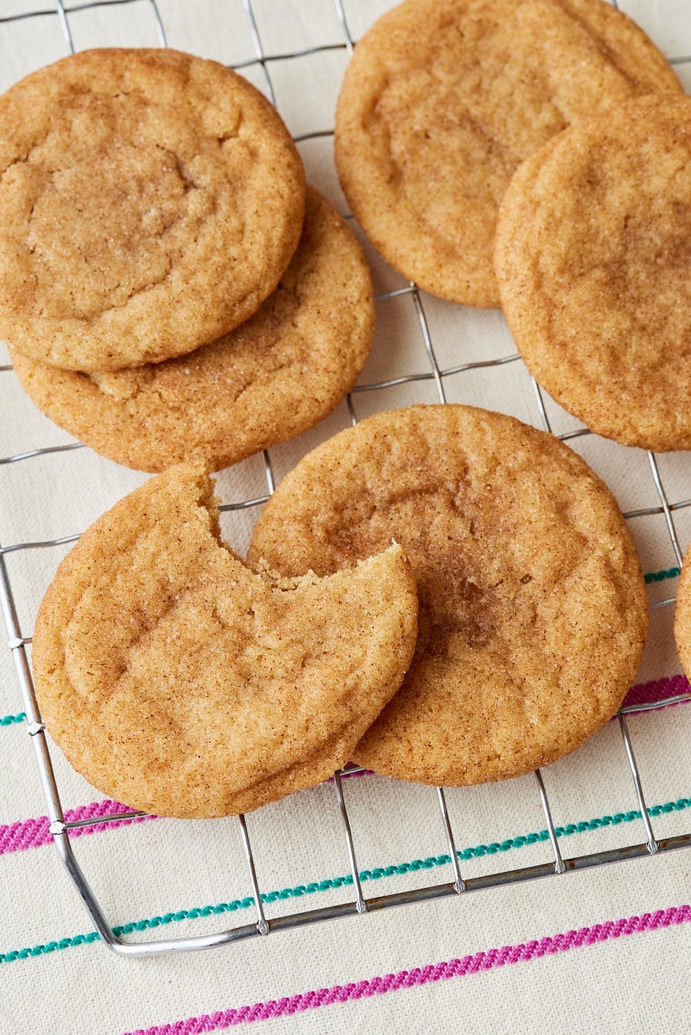 Brown Butter Snickerdoodles9 Close-up of soft, chewy brown butter snickerdoodle cookies coated in cinnamon sugar, stacked on a wire cooling rack, with one cookie broken open to reveal its tender center.