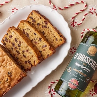 Sliced moist fruitcake loaf with raisins on a white platter, styled with Irish whiskey bottles, red and gold Christmas ornaments, and festive red-and-white twine.