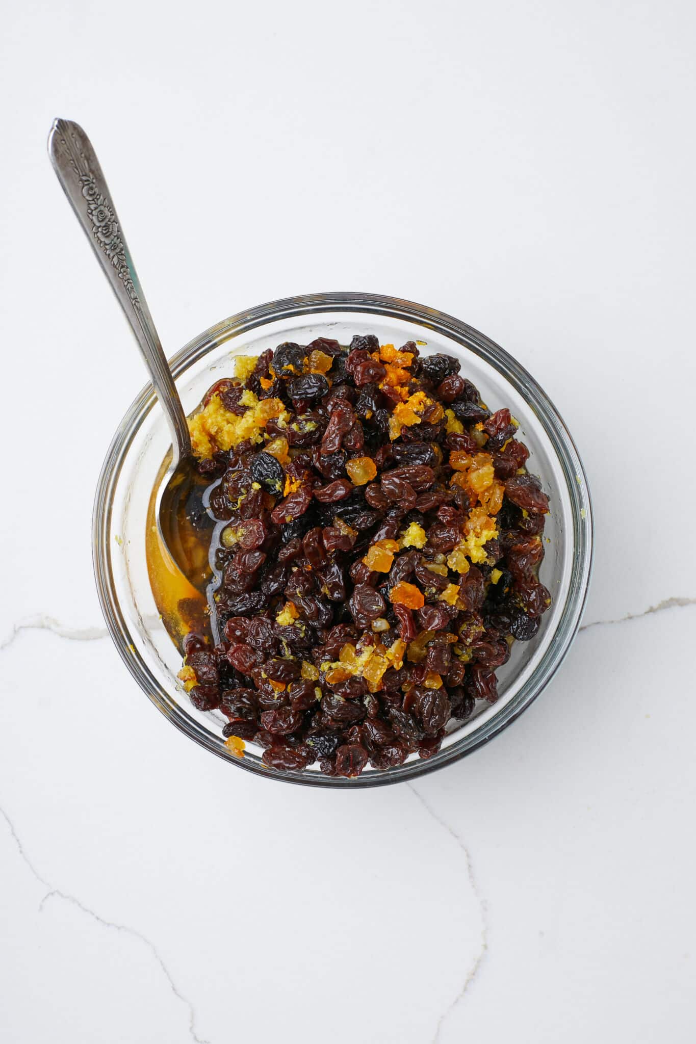 Overhead view of raisins and candied citrus peel soaking in liquid in a clear glass bowl with a metal spoon, ready for making fruitcake.