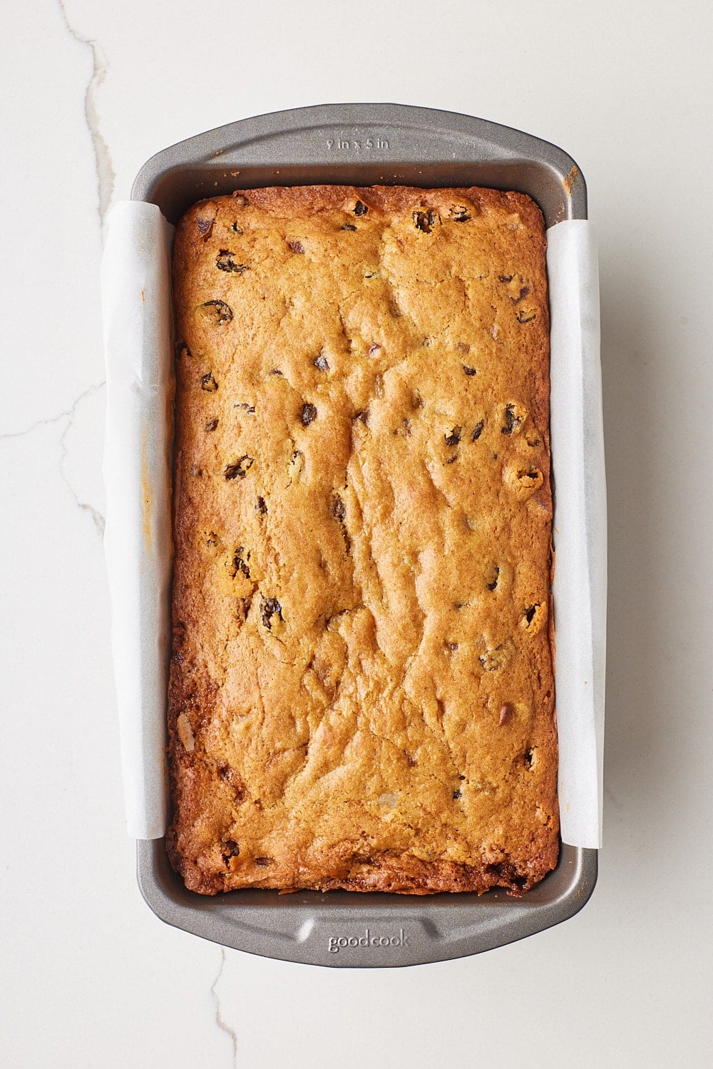 Golden-brown fruitcake loaf baked with raisins in a parchment-lined loaf pan, shown from above on a white marble countertop.