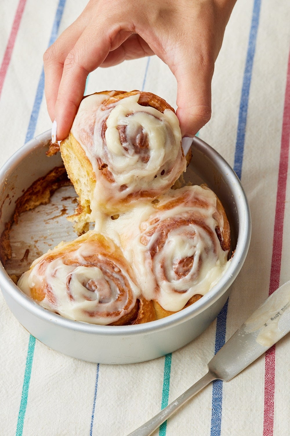Small Batch Cinnamon Rolls16 Hand lifting a freshly baked, cream-cheese-frosted cinnamon roll from a small pan of cinnamon rolls.